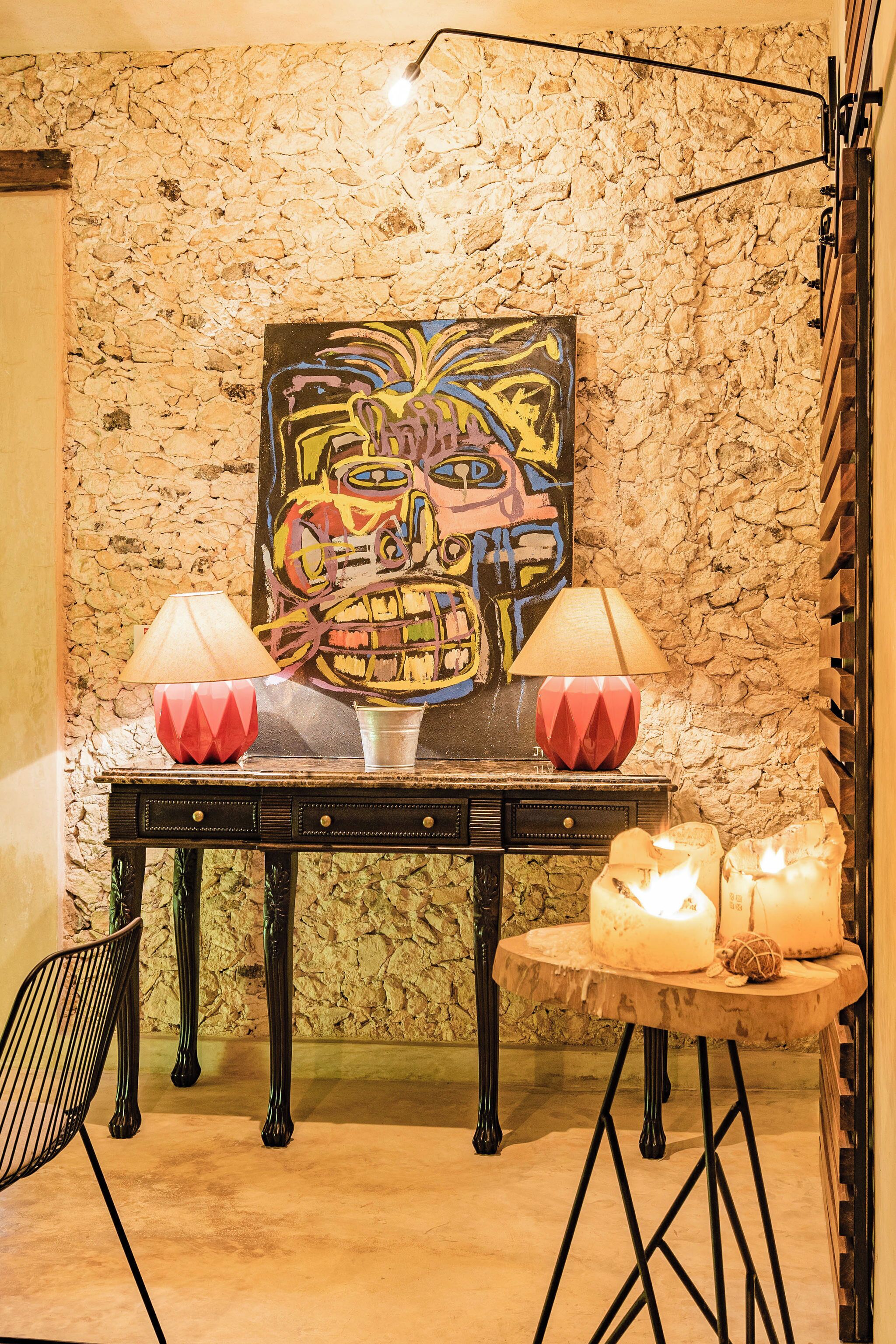 A room with a stone wall, a table with two lamps, a chair, and a painting.