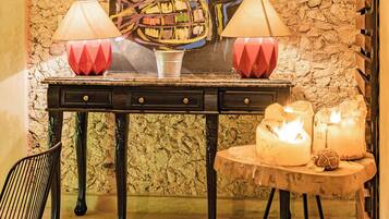 A room with a stone wall, a table with two lamps, a chair, and a painting.