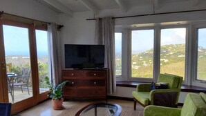 A room with a large window, a television on a wooden cabinet, a glass coffee table, and two green armchairs. - 3 Minutes Drive to Famous Magens Bay Beach (St Thomas)
