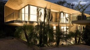 Una casa moderna con ventanas amplias y techo inclinado, rodeada por un camino de grava y pequeños árboles.
