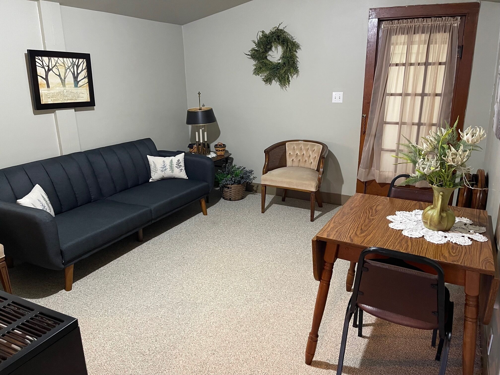 A living room with a dark blue sofa, a wooden dining table with a vase of flowers, a wooden chair, a small side table with a lamp, and a wreath on the wall.