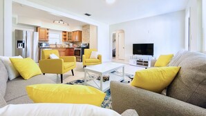 Una sala moderna con un sofá, sillones y una mesa de centro. Se ve una cocina al fondo con electrodomésticos y armarios.