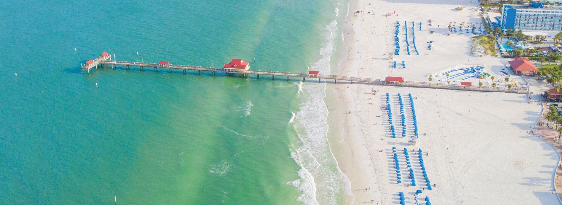 An aerial view of a beach with a pier extending into the water, rows of beach umbrellas, and a hotel with multiple floors.