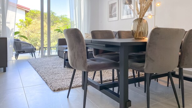 A dining area with a dark wooden table and cushioned chairs, a glass vase with dried plants, and a view of greenery outside. - Phaedrus Living: Limnaria Luxury Flat Vine 121 (Paphos)