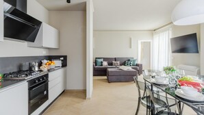 Eine moderne Küche mit Gasherd, Backofen und weißen Schränken. Es gibt einen Essbereich mit Glastisch und Stühlen sowie ein Wohnzimmer mit einer Couch und Kissen.