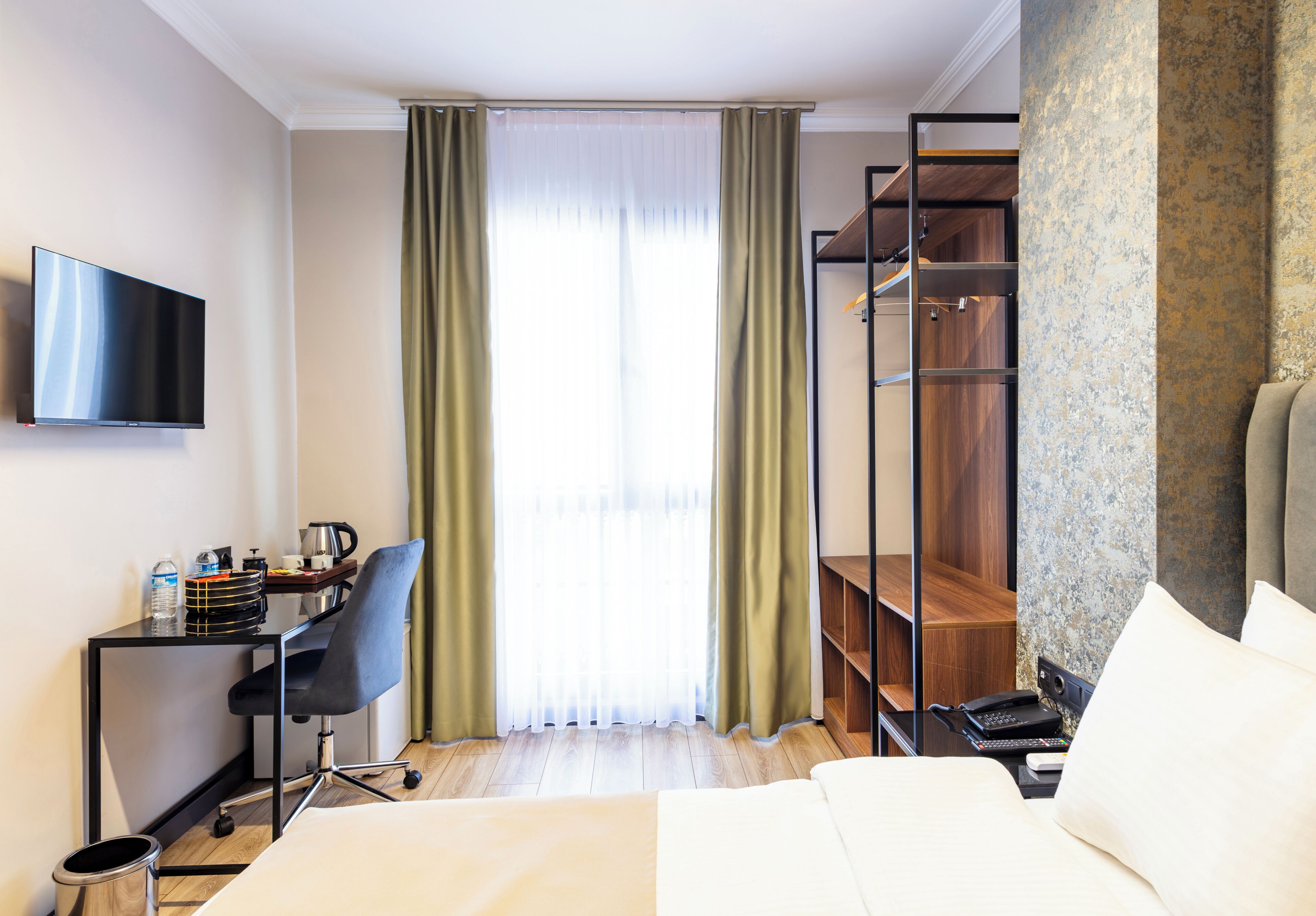 A hotel room with a bed, a desk with a chair, a TV, a wardrobe, and a window with curtains.