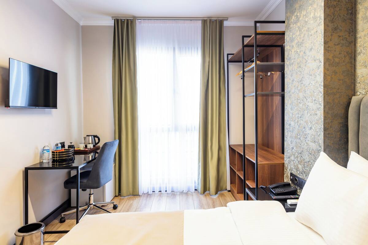 A hotel room with a bed, a desk with a chair, a TV, a wardrobe, and a window with curtains.
