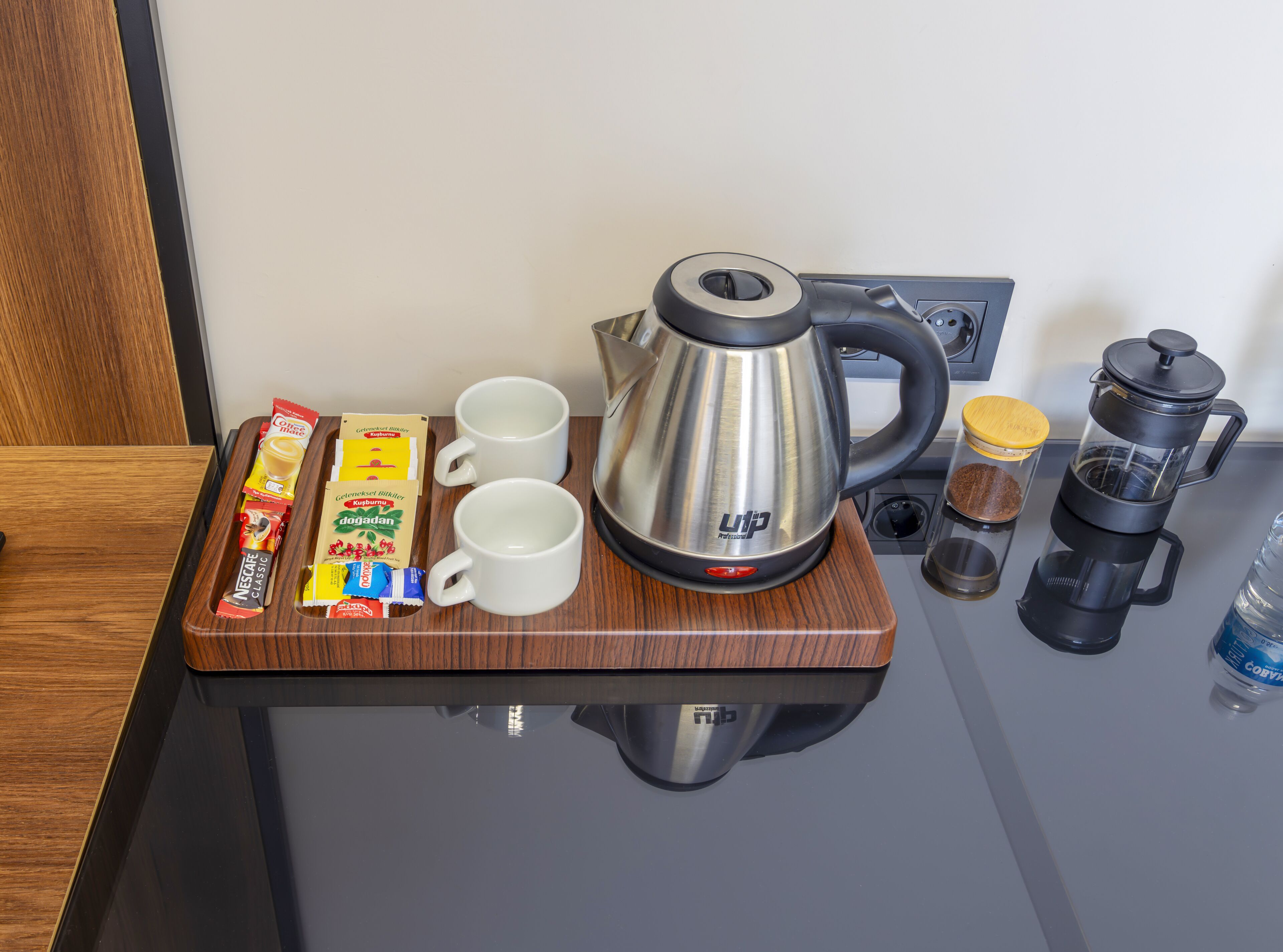 A coffee station with a coffee maker, two cups, a tray with coffee packets, a spice container, and a water bottle.