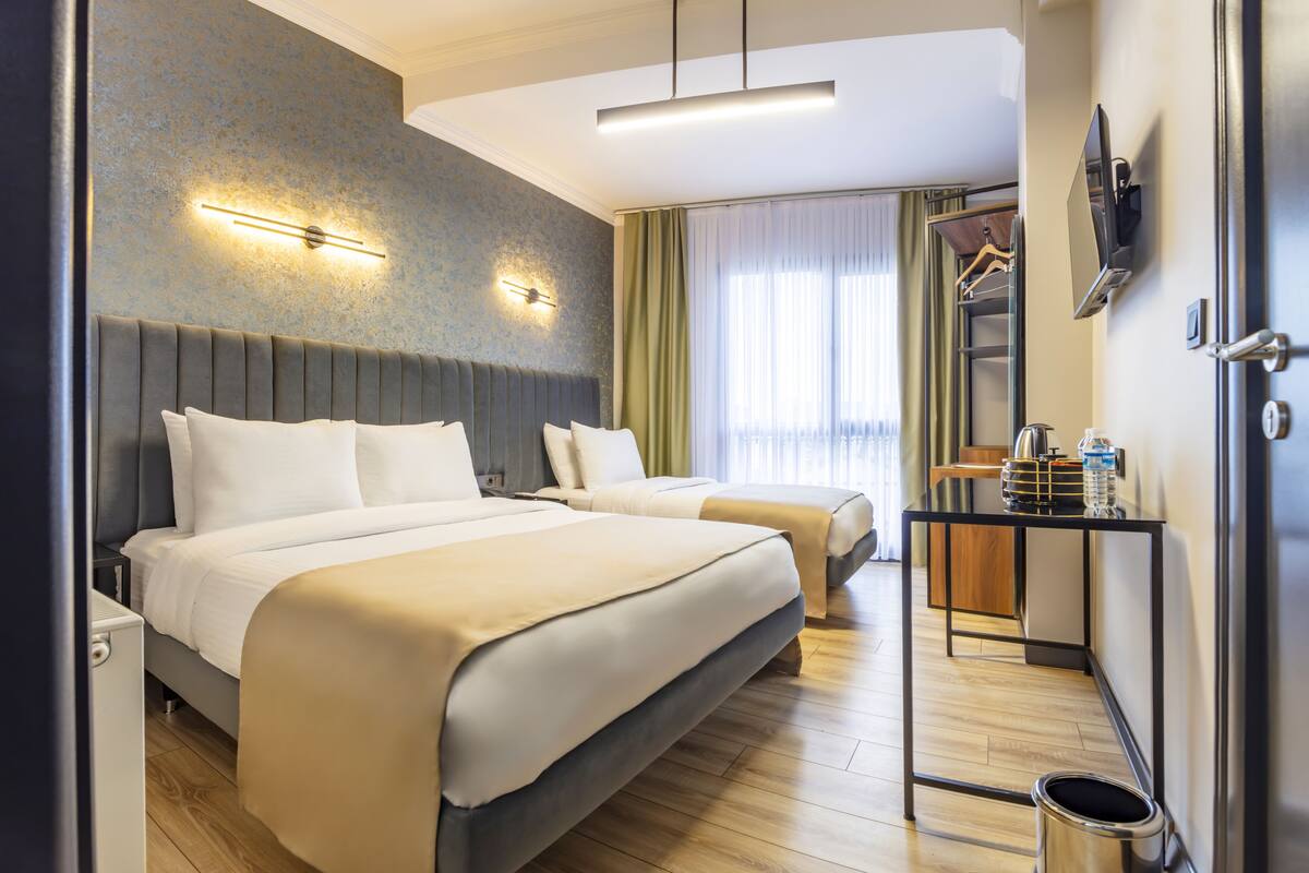 A hotel room with two beds, a bedside table, a mirror, a TV, and a window with curtains.