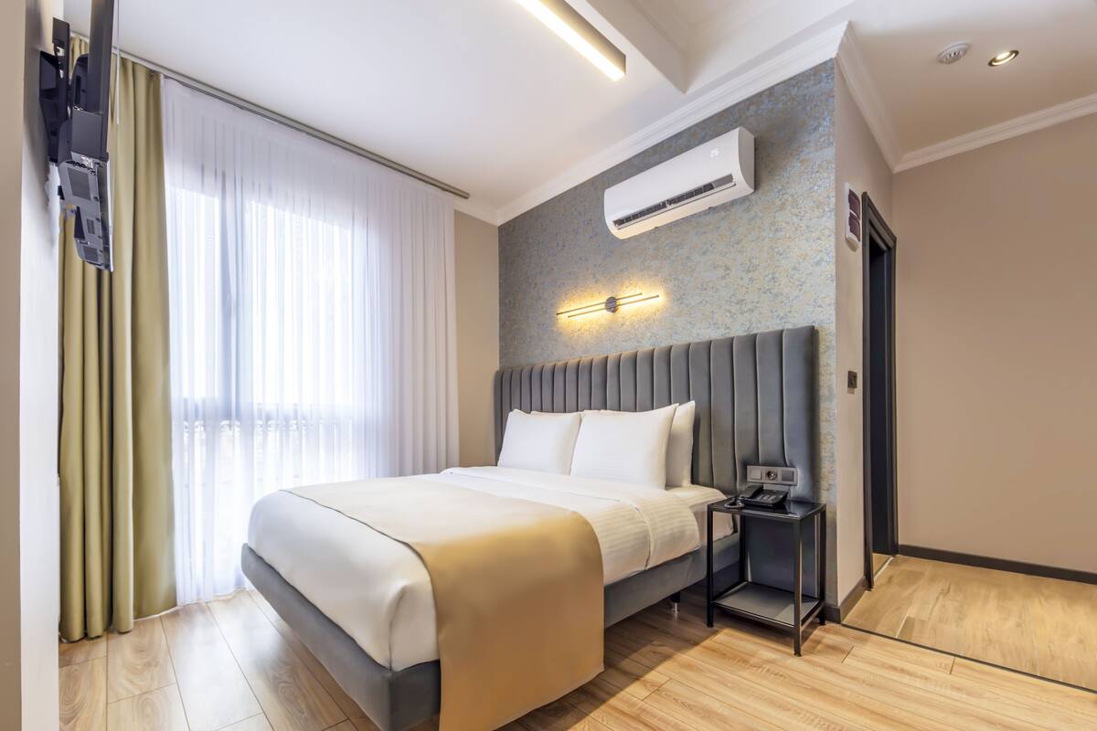A modern hotel room with a large bed, a bedside table with a phone, an air conditioning unit, and a window with curtains.