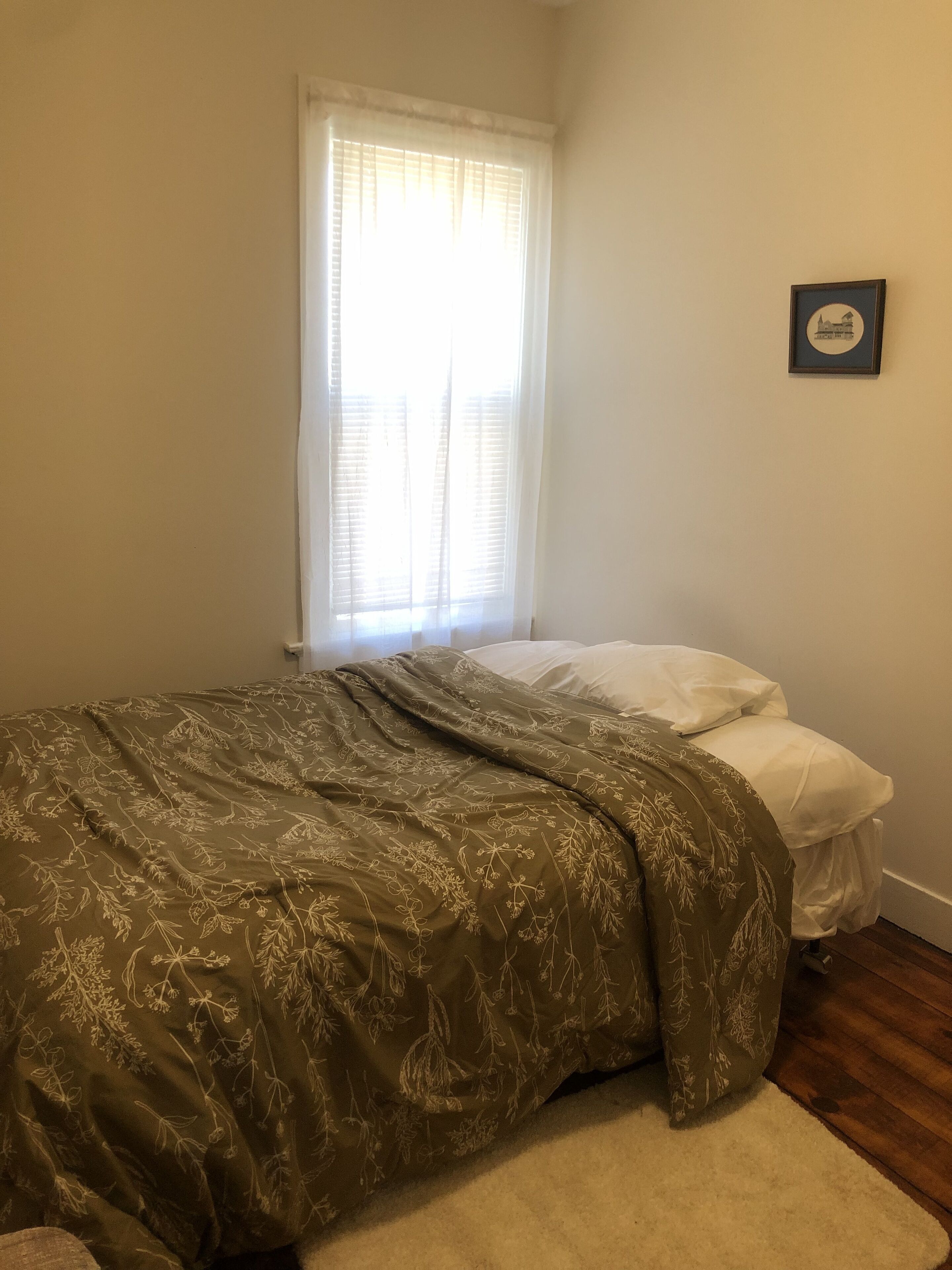 A bedroom with a bed, a window with blinds, a framed picture on the wall, and a wooden floor.