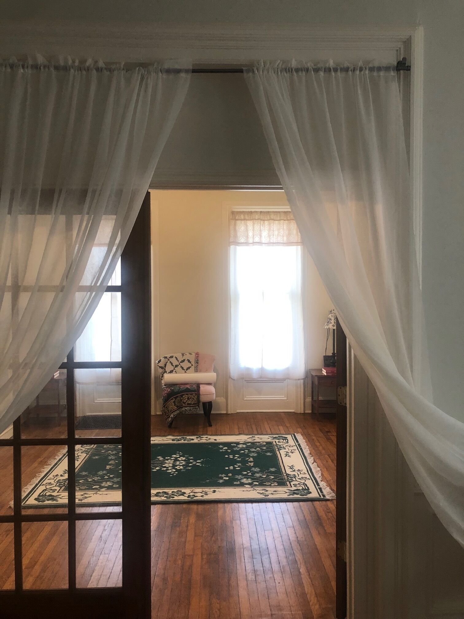 A room with a wooden floor, a patterned rug, a chair, and a window with sheer curtains.