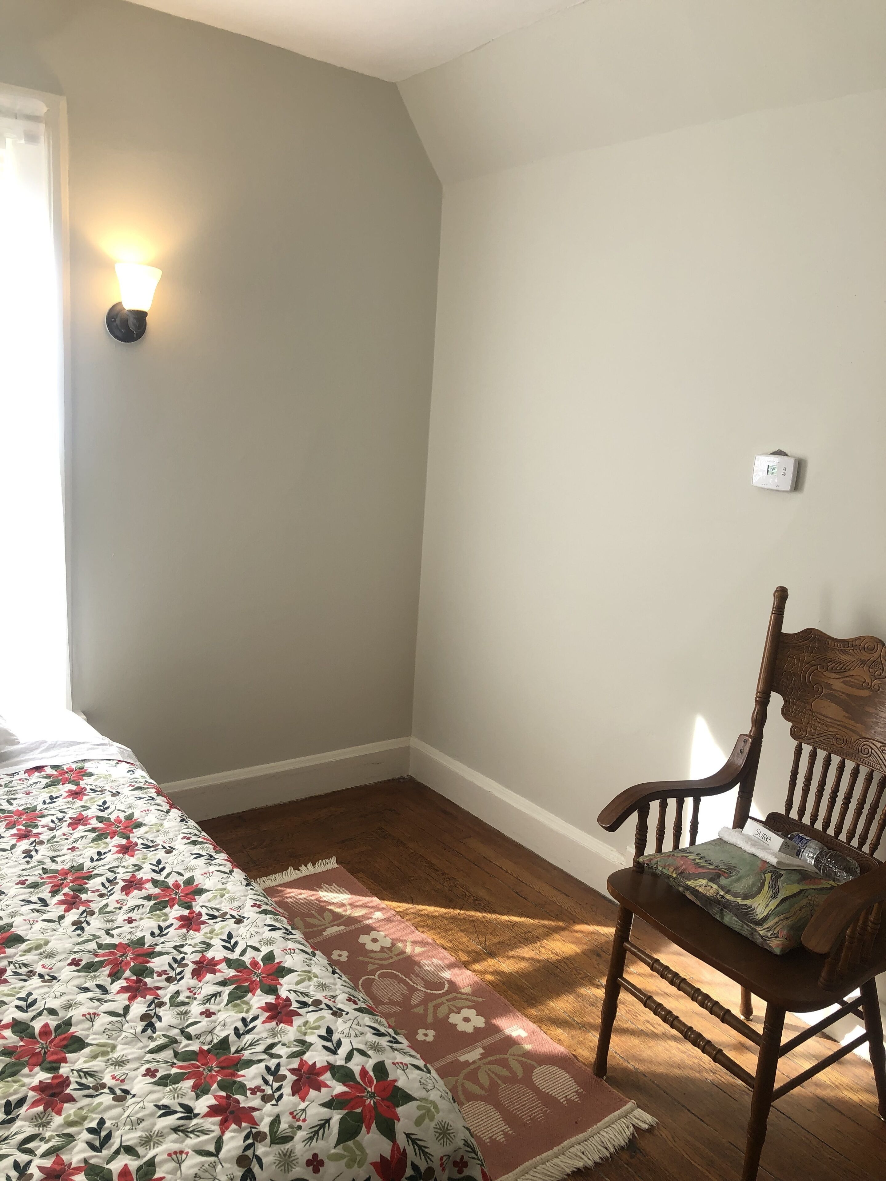 A bedroom with a bed, a chair, a wall-mounted thermostat, and a window with curtains.