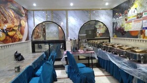 Un salón de banquetes con una larga mesa cubierta con un mantel azul, rodeada de sillas con fundas azules. Hay dos ventanas arqueadas en la pared del fondo y un mural que representa comida.