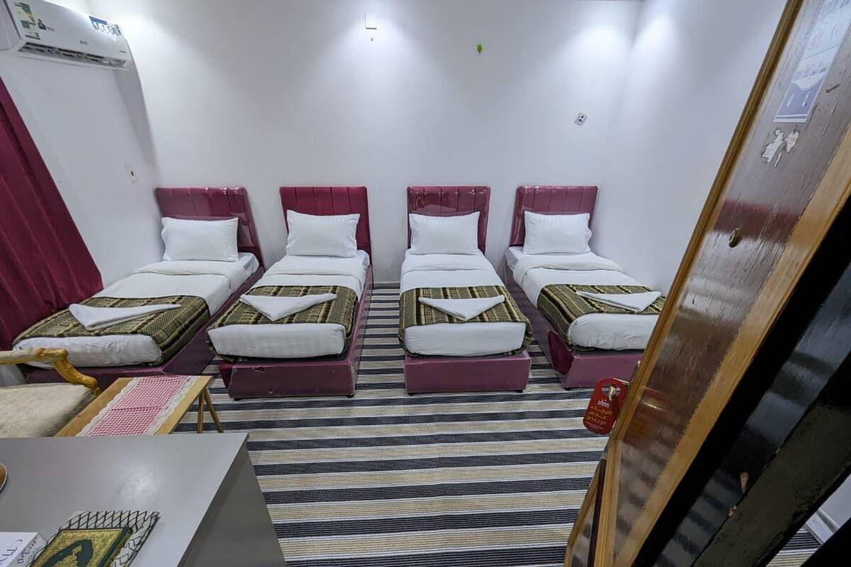 A hotel room with four beds arranged in a row, a striped rug, a small table with a book, and a wall-mounted air conditioner.