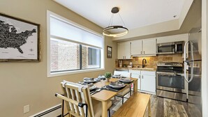 A modern kitchen with stainless steel appliances, a dining area with a wooden table and chairs, and a large window with a view of a brick building.