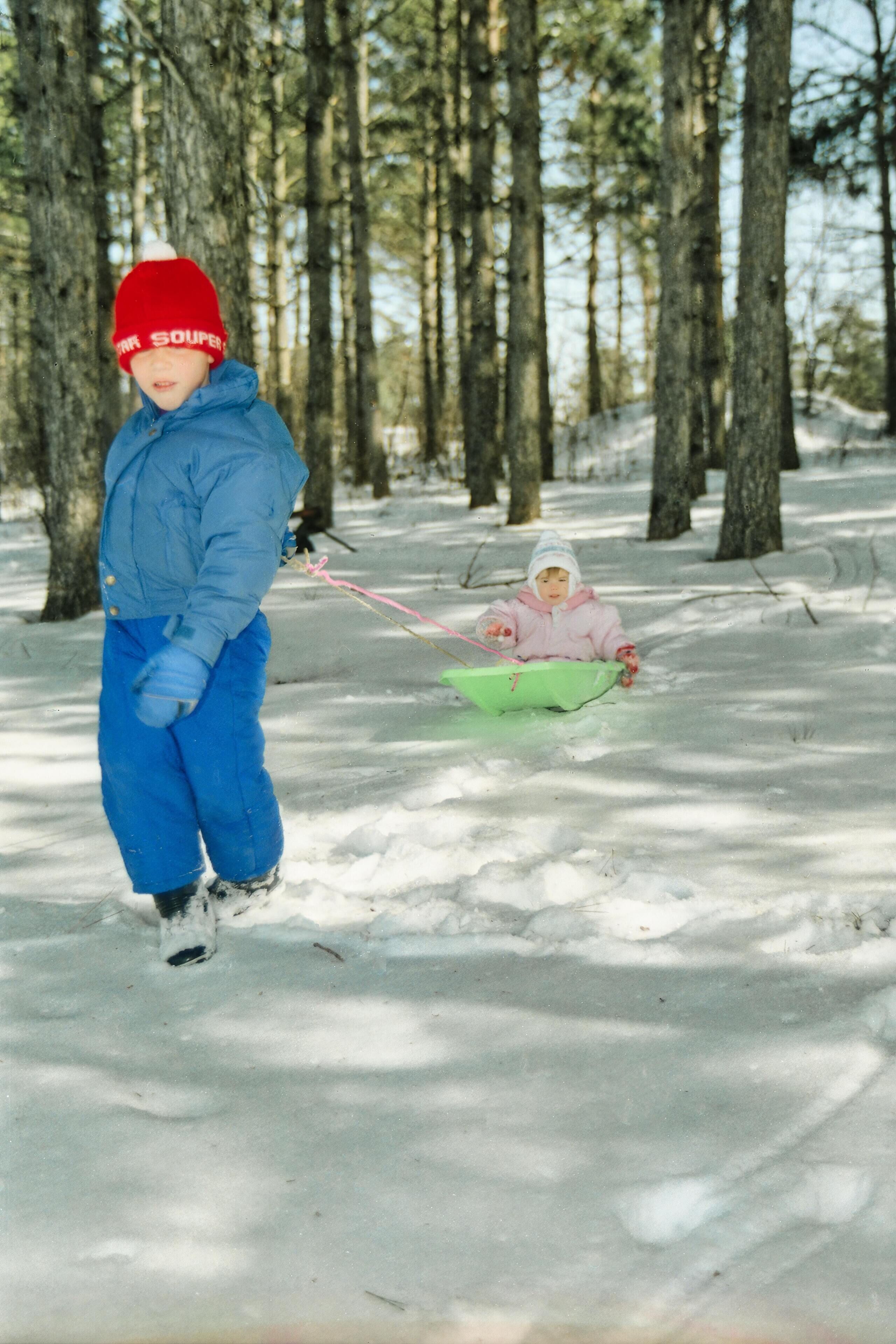 A person in a blue jacket and red hat pulling a child in a green sled through a snowy forest.