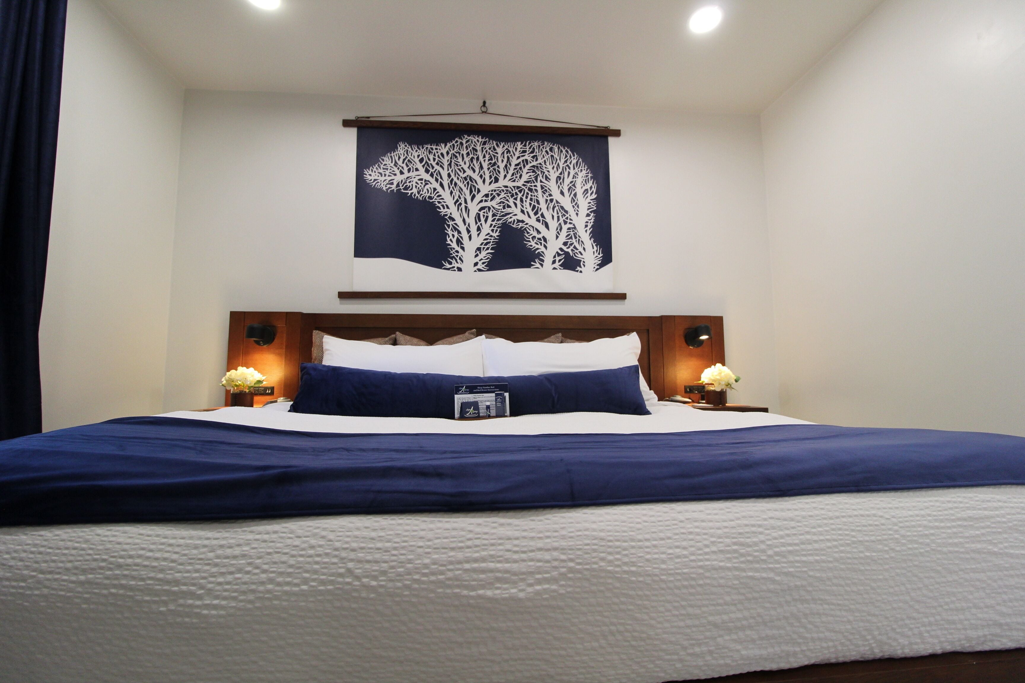 A neatly made bed with a blue and white color scheme, a wall-mounted artwork of a tree, and bedside lamps.