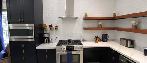 Een moderne keuken met een ingebouwde oven, magnetron en gasfornuis. Daarboven hangen houten planken en op het aanrecht staat een koffiezetapparaat.