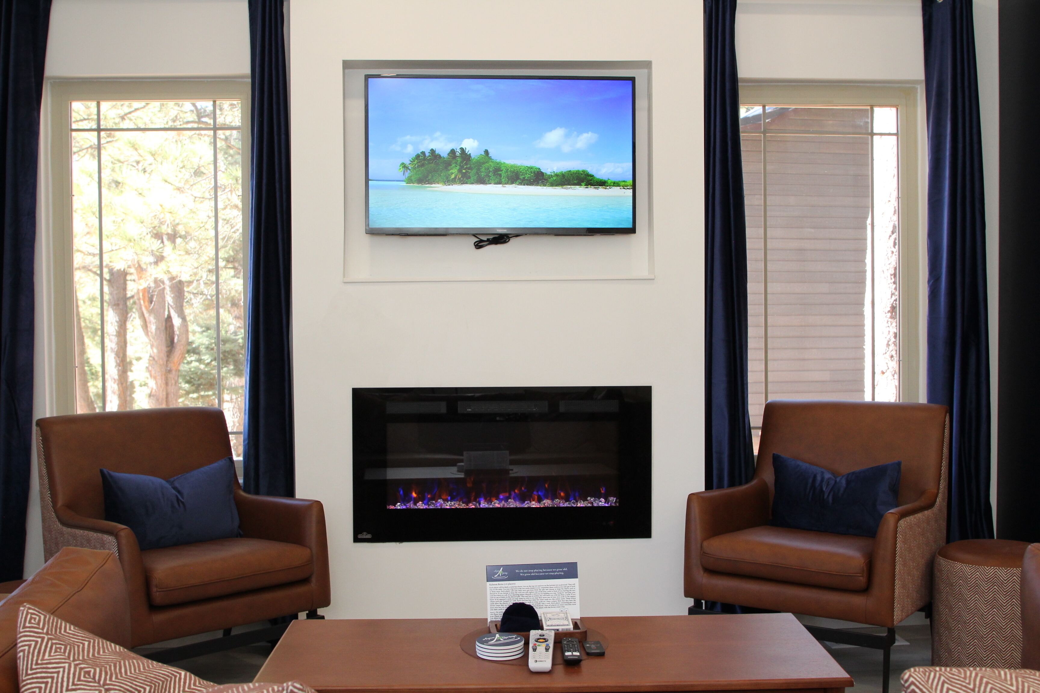 A room with a TV mounted on the wall, a fireplace, two leather armchairs, and a coffee table with remote controls.