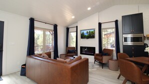 A modern living room with a brown leather sofa, a flat-screen TV mounted on the wall, and a dining area with brown chairs and a table. - Aspen Springs Angel fire (Angel Fire)