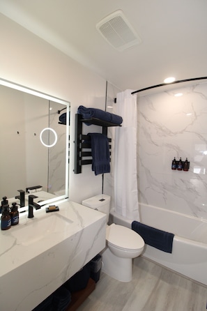 Features a sleek vanity, toilet, bathtub, and shower, with marble tiles and toiletries.
