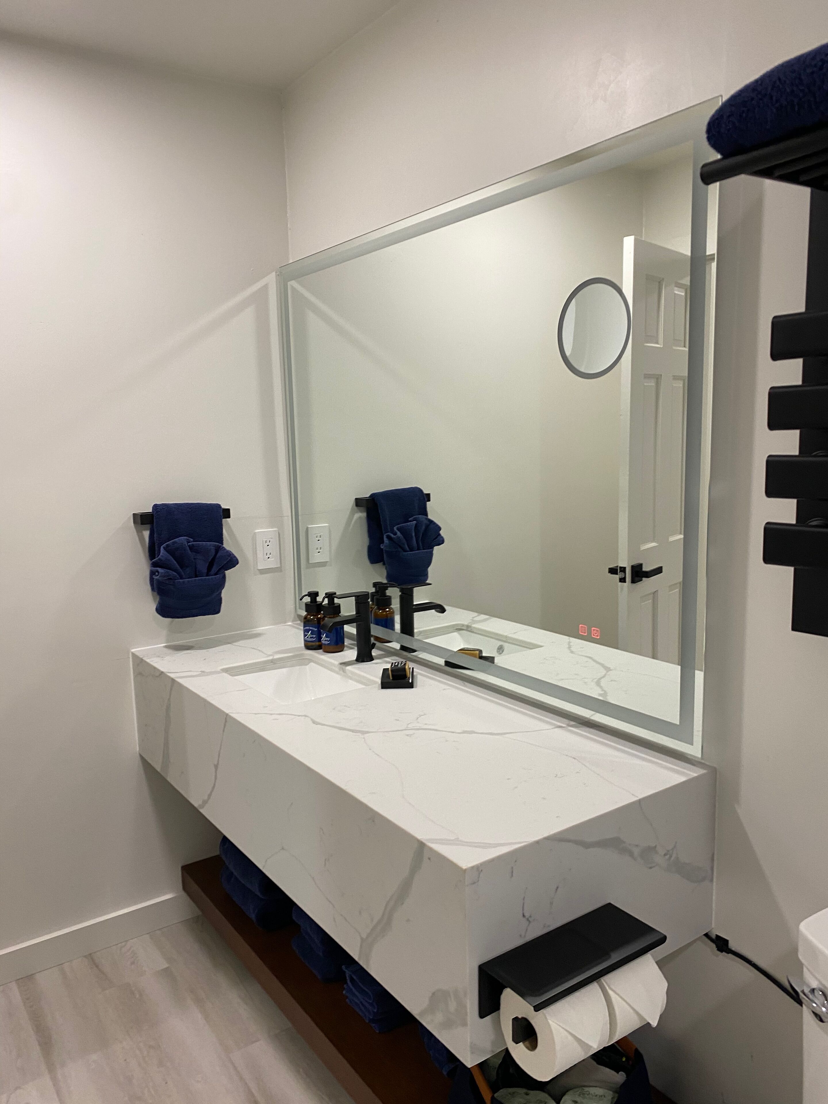 A modern bathroom with a white marble sink, two blue towels, a large mirror, and a toilet paper holder.