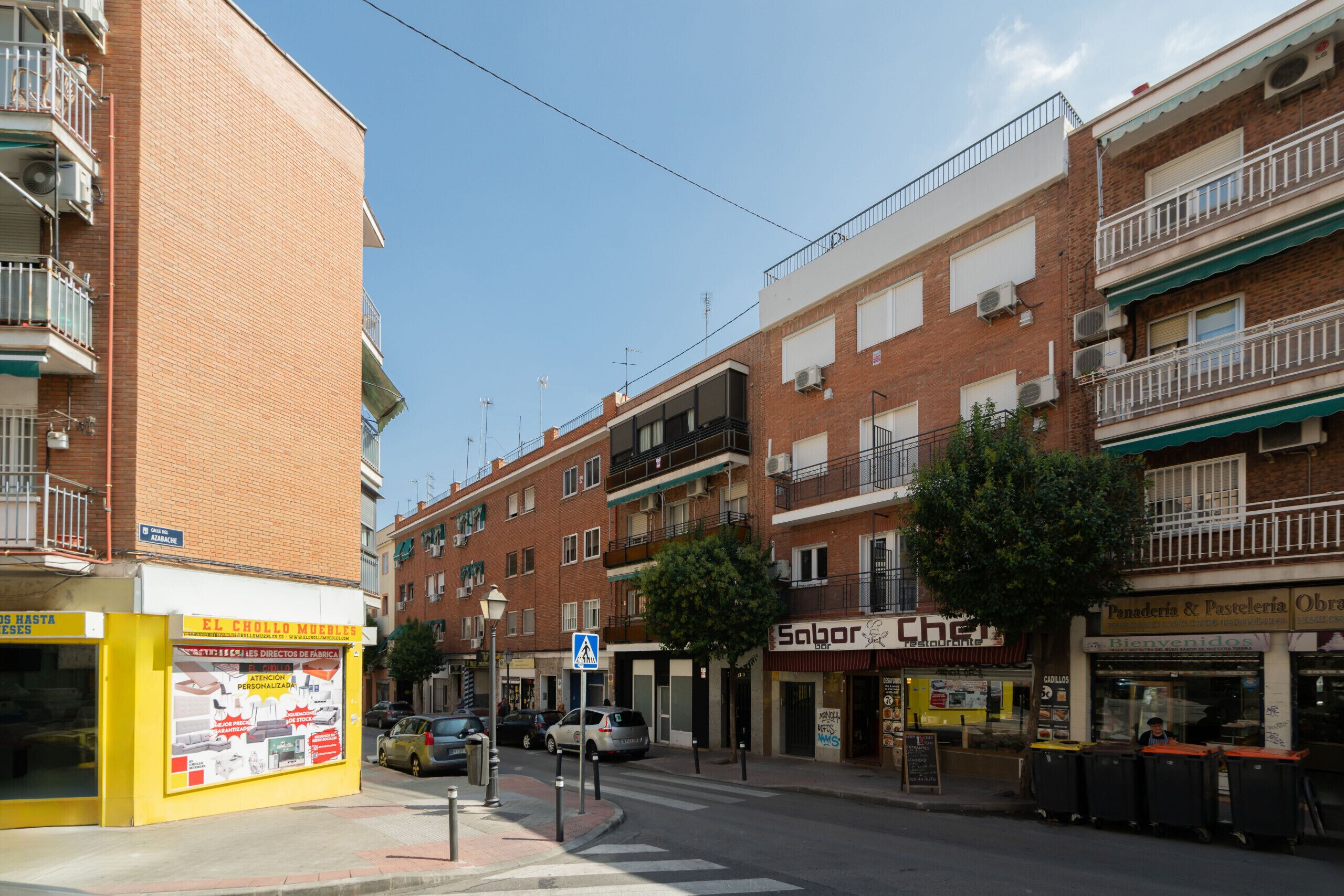 Vista da rua com uma fileira de edifícios, incluindo um restaurante chamado 'Sabor' e uma placa para 'El Chollo Muebles'.