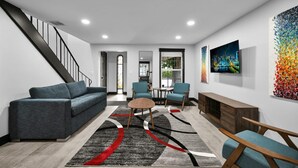 A modern living room with a grey sofa, a coffee table, and two blue armchairs. There is a staircase on the left, a large window on the right, and a colorful abstract painting on the wall. - Escape to This Gorgeous new Wilton Manors Villa (Wilton Manors)