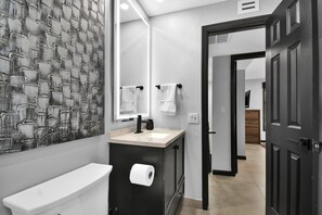 Ein modernes Badezimmer mit Toilette, Waschbecken und Spiegel. Handtücher hängen an der Wand und eine Tür führt in einen weiteren Raum.