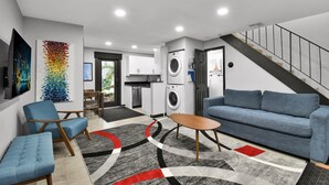 Een moderne woonkamer met een blauwe bank, een blauwe fauteuil en een houten salontafel. Achtergrond: een wasmachine en droger.