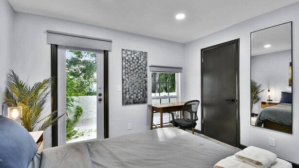 A modern hotel room with a large bed, a desk, and a chair. There is a sliding door to the outside, a large mirror, and a wall-mounted artwork.