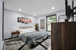 A modern bedroom with a large bed, a nightstand, a wall-mounted TV, and a window with a view of greenery. - Luxury 2-bedroom Villa in Wilton Manors (Wilton Manors)
