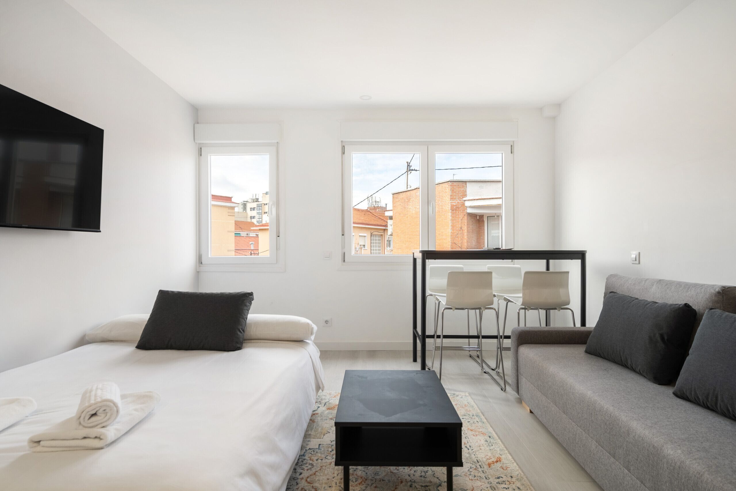 Habitación de hotel moderna con una cama, un sofá, una mesita y un televisor montado en la pared. Dos ventanas ofrecen vistas a la ciudad.
