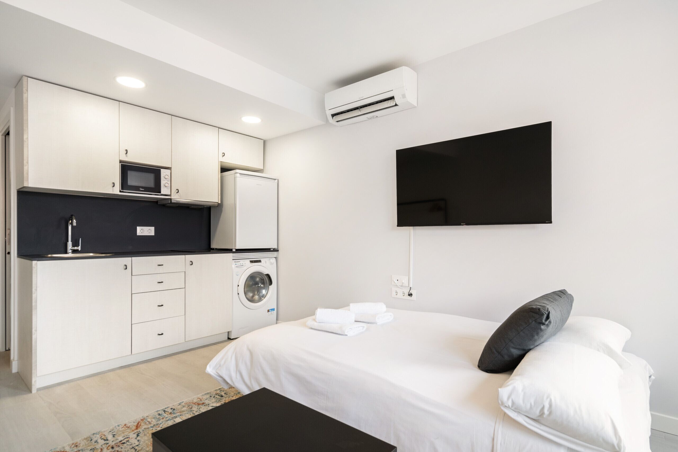 A modern hotel room with a kitchenette, a bed with white linens, a black pillow, and a mounted TV.