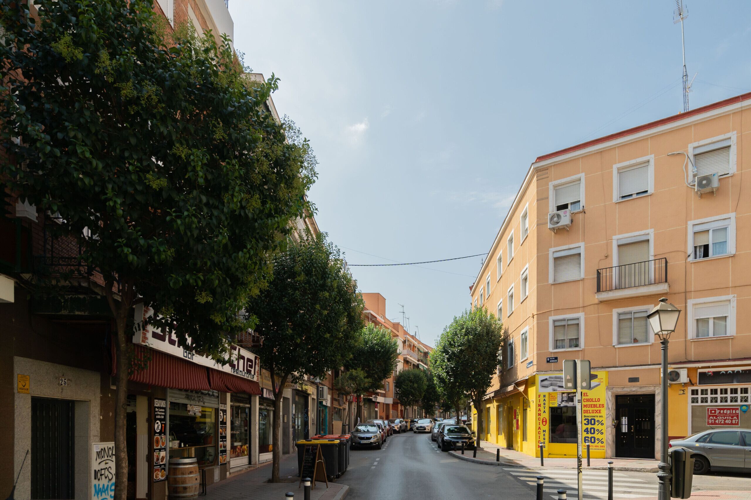 Una calle bordeada de edificios, árboles y coches aparcados.