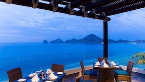 An outdoor dining area with tables set for two, overlooking a body of water and distant landforms. - Spectacular Ocean View Suite for the Holidays on the Beach!  (Cabo San Lucas)
