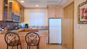 A compact kitchen with wooden cabinets, a microwave, a refrigerator, and a window. There are two chairs and a table in the center. - Spectacular Ocean View Suite for the Holidays on the Beach!  (Cabo San Lucas)