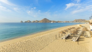 A beach with rows of sun loungers and umbrellas, a clear blue sea, and distant rocky formations. - Spectacular Ocean View Suite for the Holidays on the Beach!  (Cabo San Lucas)