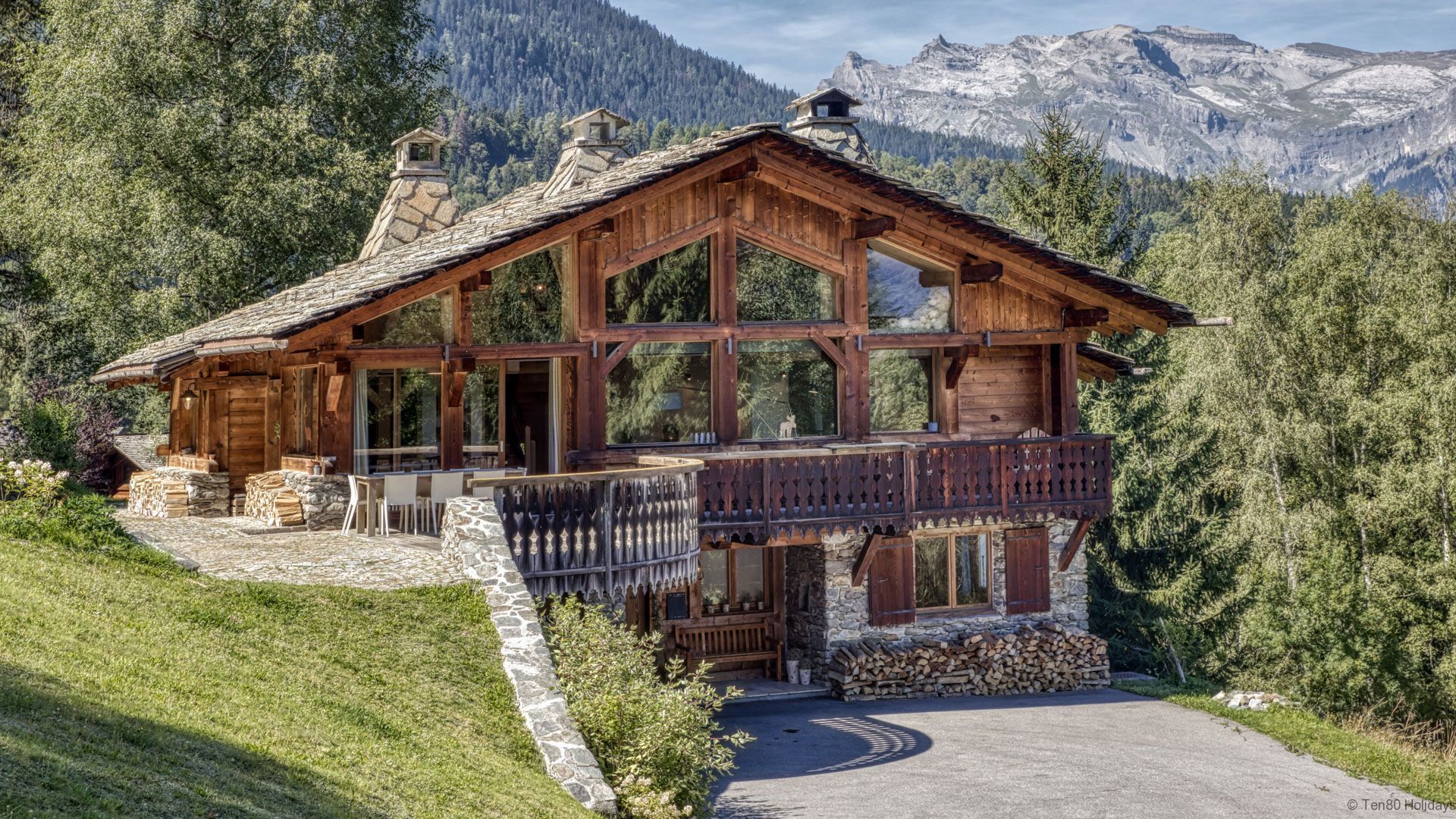 Un chalet en bois, doté de fondations en pierre, de grandes fenêtres et d’un balcon offrant une vue sur un paysage montagneux.