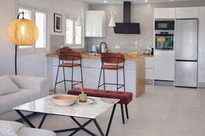 Una cocina moderna con armarios blancos, una campana extractora negra y electrodomésticos de acero inoxidable. Hay una zona de comedor con una mesa de mármol y dos sillas.