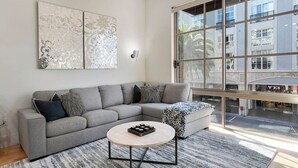 Una sala moderna con un sofá seccional gris, una mesa de centro redonda y ventanas amplias con vistas a un edificio y palmeras.