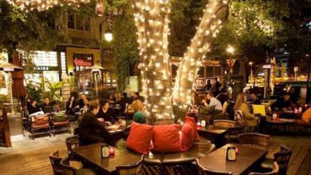 Un área de comedor al aire libre con mesas y sillas de madera, rodeada de follaje y luces colgantes.