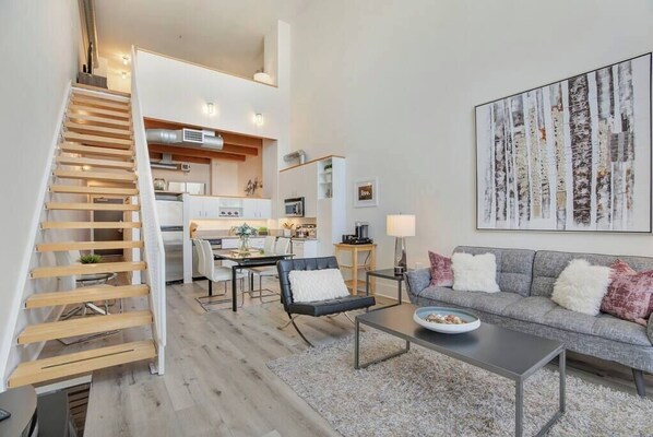 A modern apartment with a staircase leading to a kitchen, a living area with a sofa and coffee table, and a dining area with chairs.