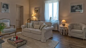 Un salon comprenant un canapé, deux fauteuils, une table basse et une petite table d’appoint avec une lampe. Il y a une fenêtre avec des rideaux légers, un radiateur et des tableaux encadrés accrochés aux murs.