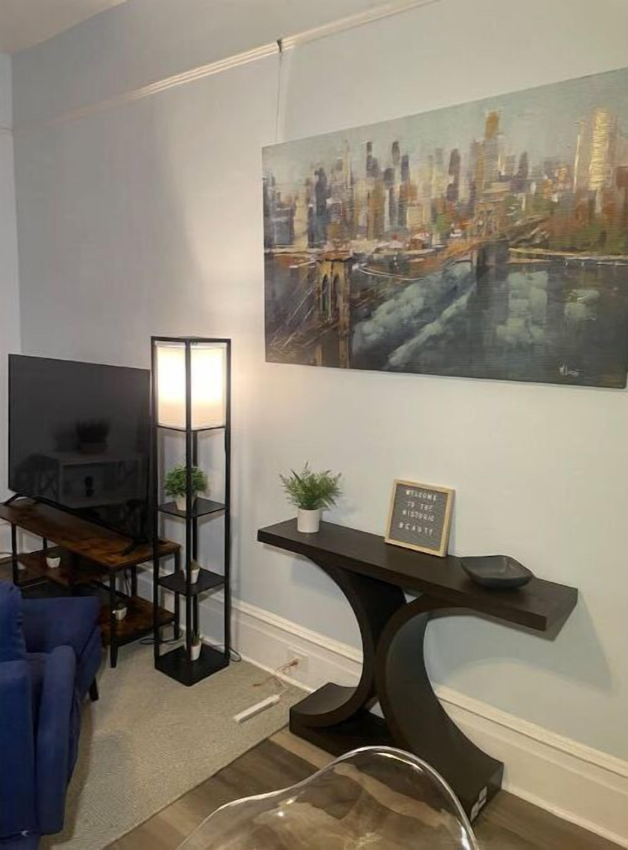 Een woonkamer met een televisie, een vloerlamp, een console met een plant en een schilderij van een stadsgezicht.
