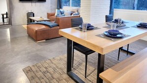 Una sala de estar moderna con un televisor de pantalla plana grande montado en la pared, una mesa de comedor de madera dispuesta con platos y vasos, un sofá de cuero con cojines azules y un amplio ventanal con vistas a árboles.