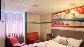 A hotel room with a bed, a red chair, a nightstand, and a wall-mounted shelf with red spherical objects.