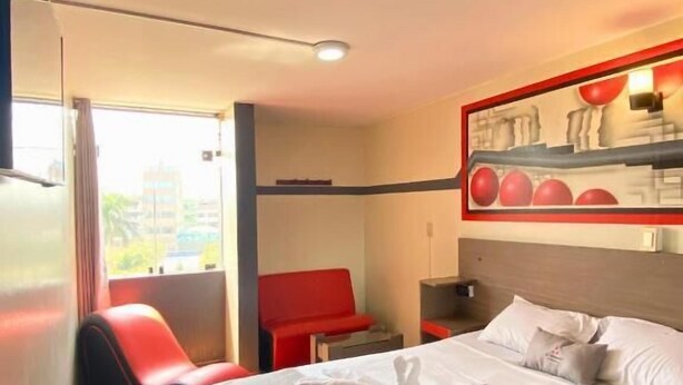 A hotel room with a large bed, a red chair, a small table, and a wall-mounted shelf with decorative items.