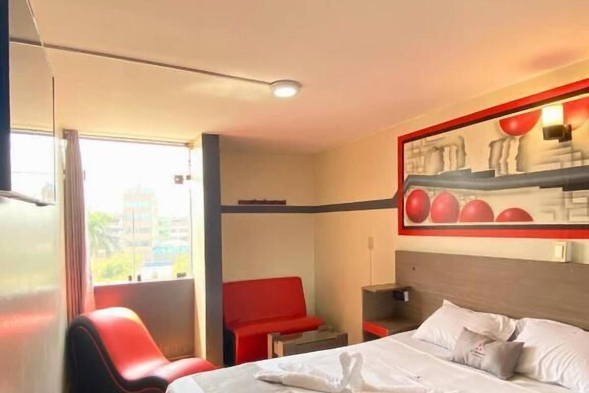 A hotel room with a large bed, a red chair, a small table, and a wall-mounted shelf with decorative items.